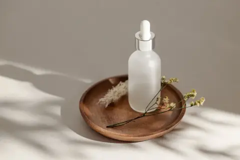 Szklana buteleczka z pipetką na drewnianej tacy, ozdobiona suchymi kwiatami. Uosobienie slow beauty, spokoju i naturalności.